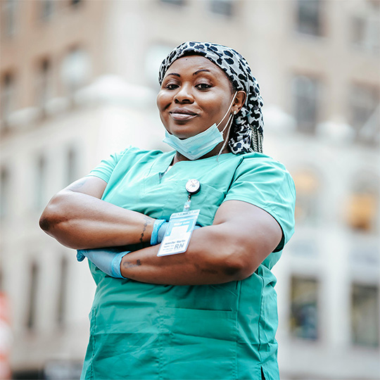 A woman wearing a scrub suit stands in the center of a street, showcasing her professional attire against a city backdrop.