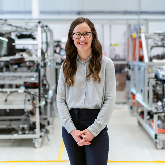 A woman wearing glasses stands confidently in front of some industrial equipment.