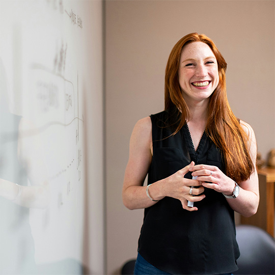 A woman smiles confidently while standing in front of a whiteboard filled with notes and diagrams.