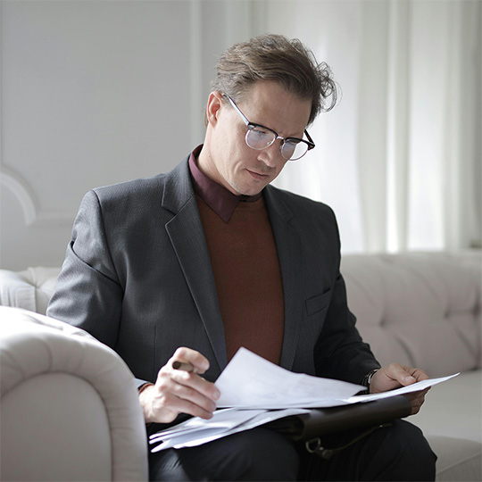 A man in business attire sits on a sofa, intently reading through important documents.