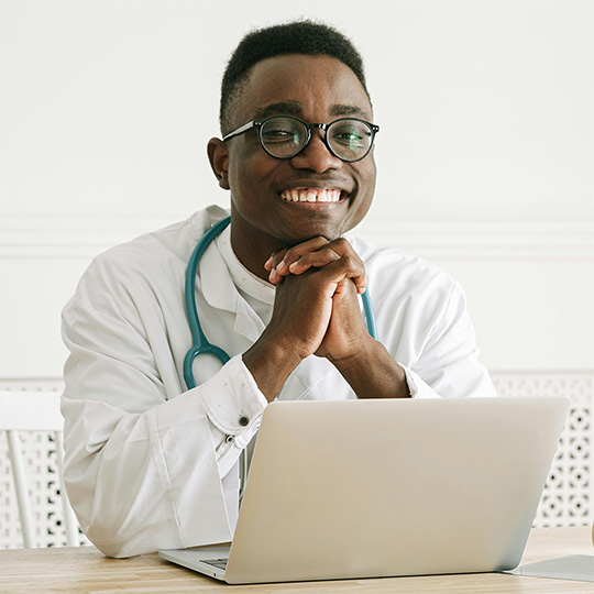  A cheerful Black man in a white lab coat is seated at a desk, engaged with his laptop, exuding professionalism.