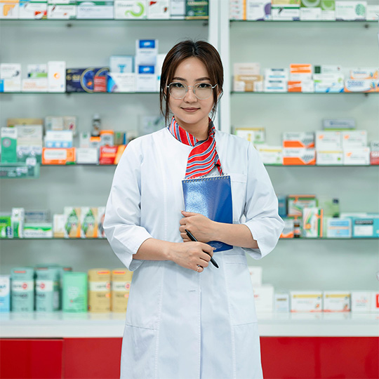 A female pharmacist stands confidently before shelves, looking directly at the camera in a modern pharmacy.