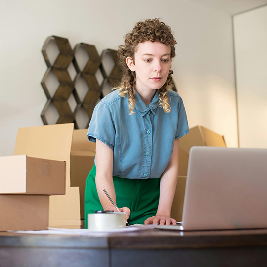 A woman typing on her laptop, with several boxes stacked around her in a workspace.