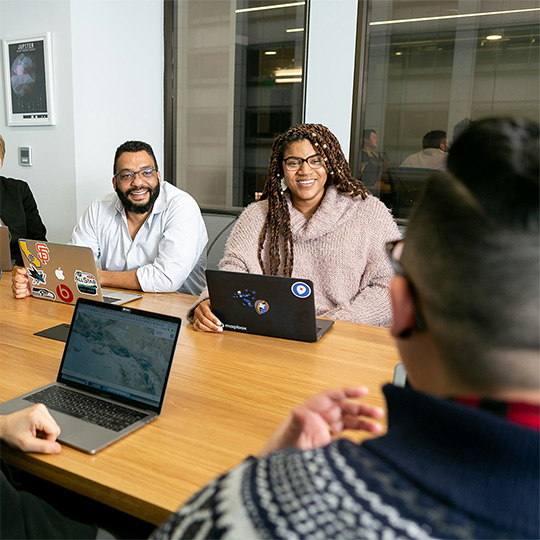 Several people engaged in a meeting, seated at a table with laptops open, focused on their work and ideas.