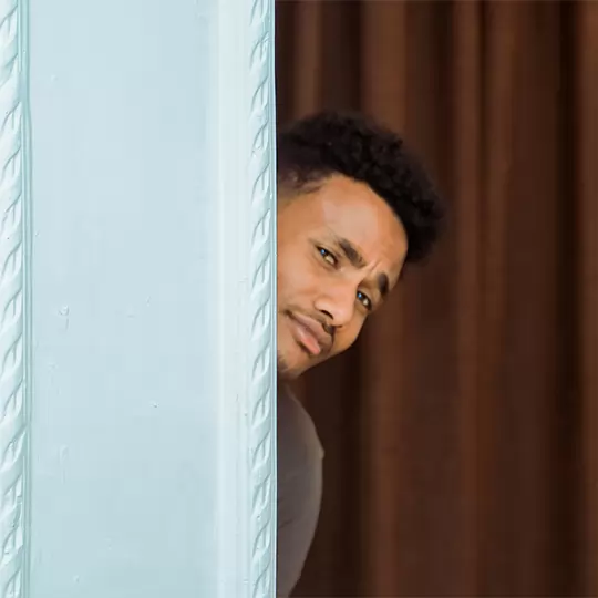 A man cautiously peeks out from behind a partially open door, revealing a curious expression on his face.